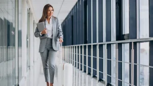 Mulher sorridente em traje de negócios cinza, com um laptop na mão, puxa uma mala de rodinhas branca por um corredor iluminado de aeroporto.