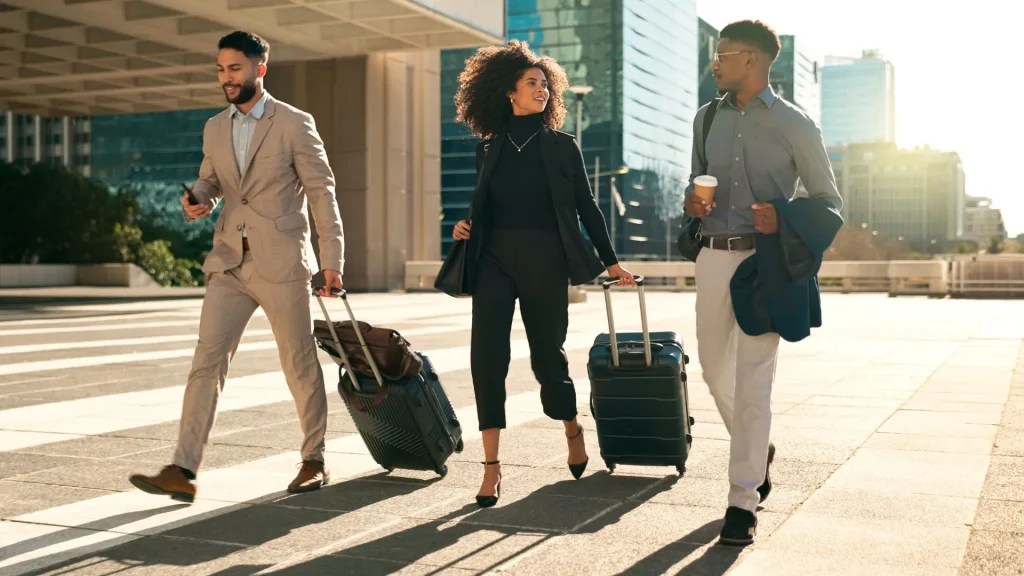 Três profissionais, dois homens e uma mulher, sorrindo e caminhando lado a lado em uma calçada de cidade ensolarada, cada um com uma mala.