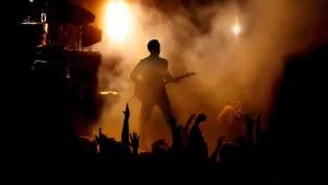 Uma silhueta de um guitarrista no palco, com luzes brilhantes e fumaça ao fundo, enquanto as mãos da multidão se erguem em adoração. A iluminação laranja e amarela cria uma atmosfera energética.