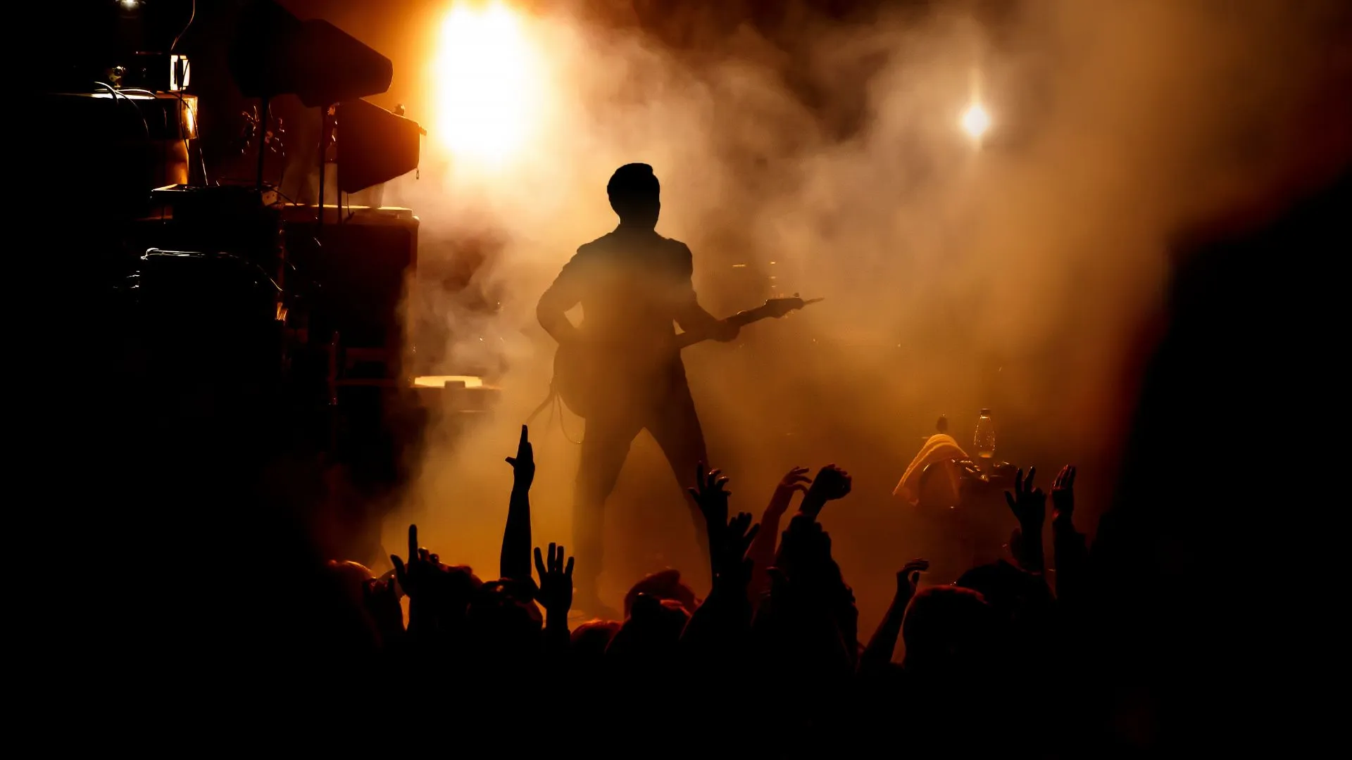 Uma silhueta de um guitarrista no palco, com luzes brilhantes e fumaça ao fundo, enquanto as mãos da multidão se erguem em adoração. A iluminação laranja e amarela cria uma atmosfera energética.