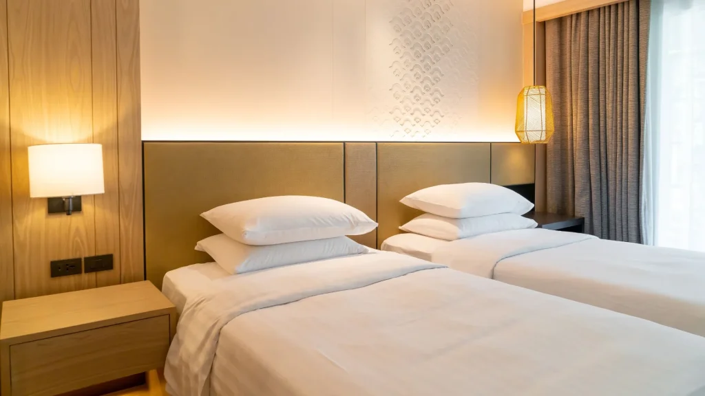 Dois leitos de solteiro com lençóis brancos e travesseiros empilhados são vistos de perto em um quarto de hotel moderno e aconchegante. A decoração tem painéis de madeira claros e uma cabeceira estofada em bege, iluminada por uma faixa de LED. Uma luminária suspensa de vime e um abajur de parede com uma base de madeira e um interruptor completam a iluminação e adicionam um toque de estilo natural.