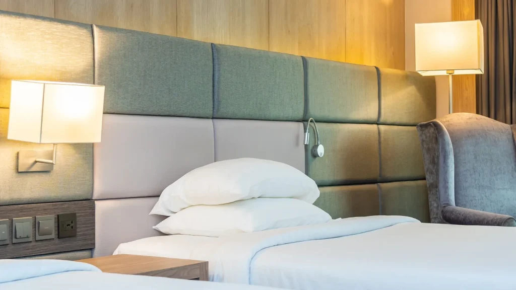 Duas camas de solteiro de um hotel com lençóis brancos e travesseiros empilhados. À esquerda, uma luminária de parede ilumina um pedaço de madeira. No centro da cabeceira cinza, há outra luminária de parede. À direita, um abajur de pé ilumina uma poltrona cinza.