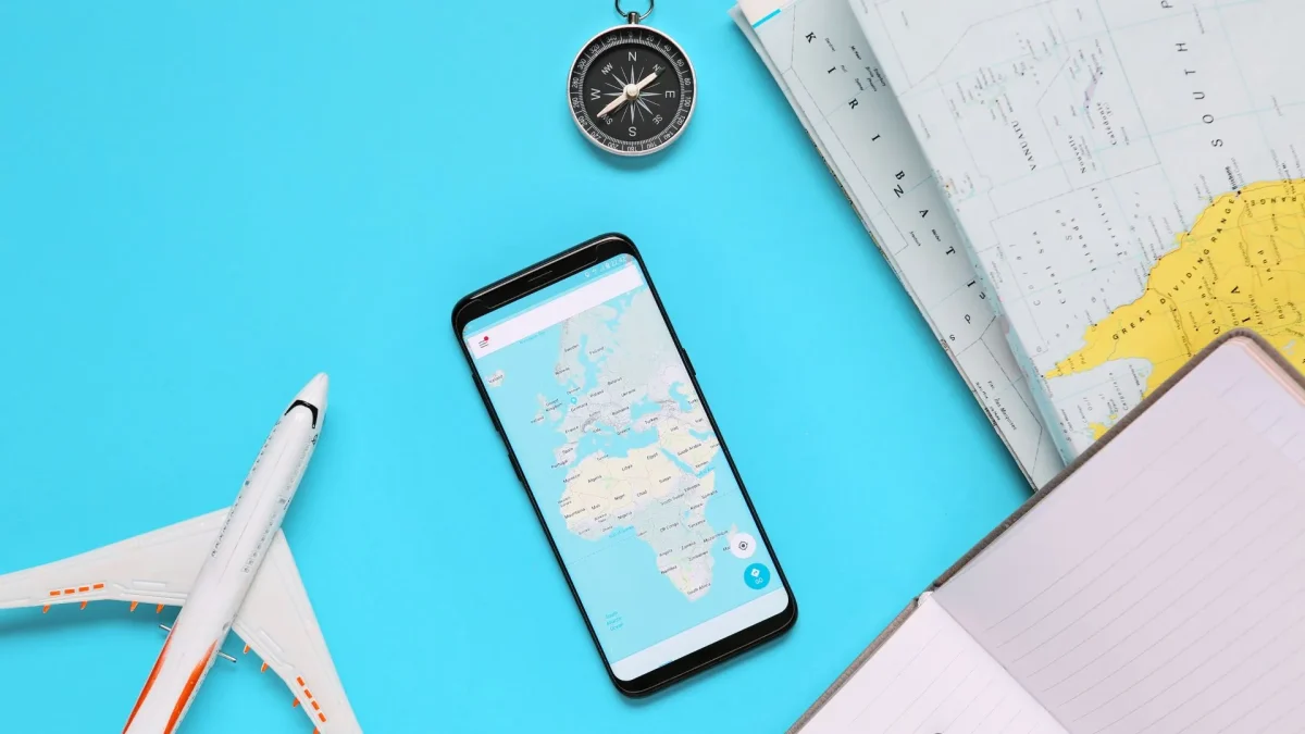 Objetos relacionados a viagens, como um pequeno avião de brinquedo, um smartphone mostrando um mapa, uma bússola e um caderno com uma caneta, dispostos sobre uma superfície azul clara. A imagem sugere planejamento ou preparação para uma viagem.