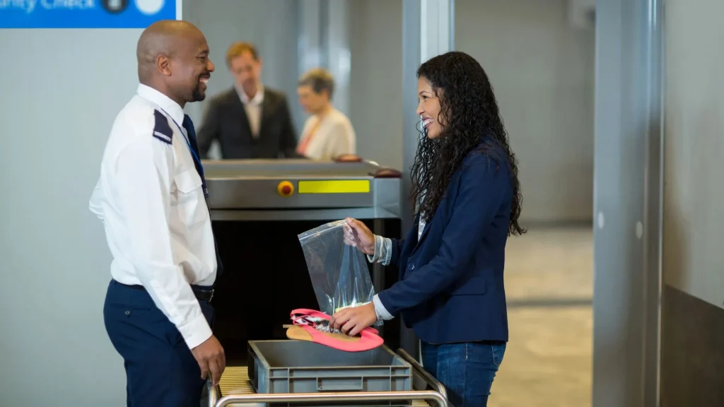 Um oficial de segurança sorridente, vestindo uniforme branco e azul, interage com uma passageira em um posto de verificação de aeroporto. A mulher, também sorrindo, segura uma sacola plástica transparente com frascos pequenos e coloca um par de sandálias cor-de-rosa em uma bandeja cinza sobre a esteira do raio-X. Ao fundo, outros passageiros aguardam na fila.