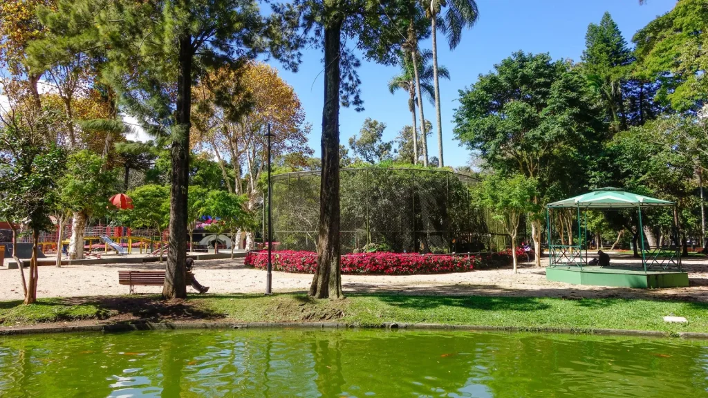 Vista de uma área do Passeio Público de Curitiba, no Paraná. Em primeiro plano, um lago de água verde. Ao centro, um grande viveiro de pássaros cercado por flores vermelhas. Do lado direito, um coreto verde com telhado. Do lado esquerdo, um parquinho infantil com balanços. A cena é cercada por diversas árvores.