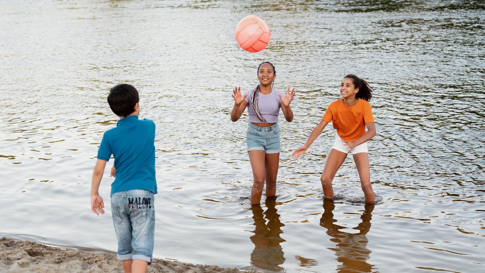 Três crianças brincam em um rio. Duas meninas, de pé na água até a altura das canelas, estão no centro e na direita da imagem, e um menino, de pé na areia, está de costas para o observador na parte esquerda. A menina no centro, que veste uma blusa roxa, joga uma bola rosa para o alto, em direção ao menino. A segunda menina, de blusa laranja, está à direita e parece pronta para a jogada. O rio tem uma superfície com pequenas ondulações. O fundo da imagem está desfocado, mostrando a água em tom acinzentado.