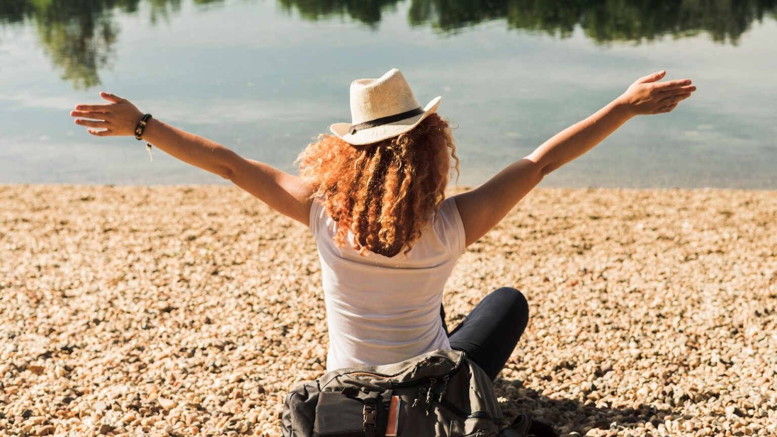 Uma mulher de chapéu de palha e cabelo ruivo e cacheado, sentada de costas para a câmera, de frente para um lago. Ela está com os braços abertos, em um gesto que sugere liberdade ou felicidade. Ela está sentada na margem de um lago, em uma área coberta por pequenas pedras e areia. A mulher usa uma blusa branca e calça escura, e há uma mochila escura na frente dela.