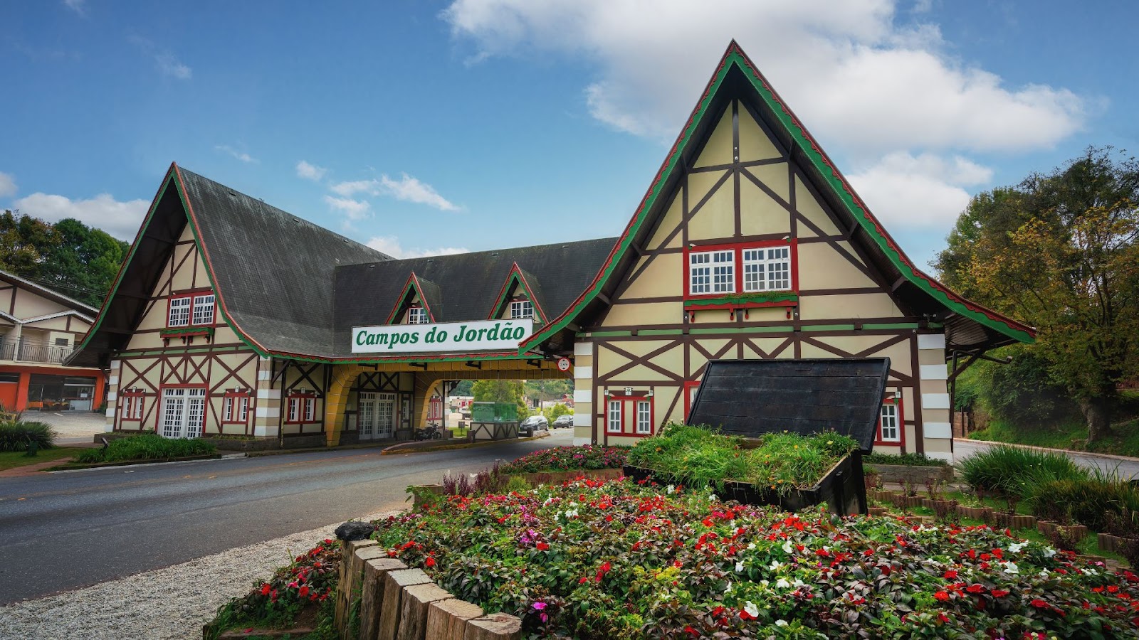 Uma fotografia diurna do portal de entrada de Campos do Jordão, em estilo arquitetônico suíço-alpino. A estrutura tem telhados íngremes e é decorada com detalhes em madeira escura sobre paredes claras. Há uma placa verde com o nome "Campos do Jordão" no arco central. Na frente do portal, há canteiros de flores coloridas e grama, e a imagem é capturada em um dia com céu parcialmente nublado.