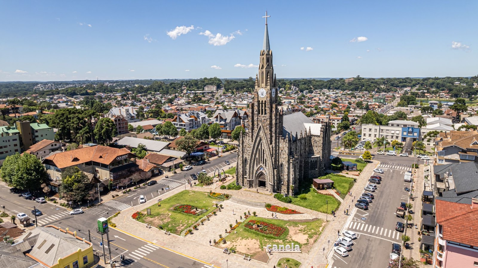 Uma visão aérea de uma cidade, mostrando uma majestosa igreja de estilo gótico com uma torre alta e pontiaguda no centro. A igreja, feita de pedra escura, está em frente a uma praça verde com canteiros de flores. A cidade ao redor é composta por prédios de telhados vermelhos e outras construções, cercadas por muitas árvores. O céu está azul e com poucas nuvens, indicando um dia ensolarado.