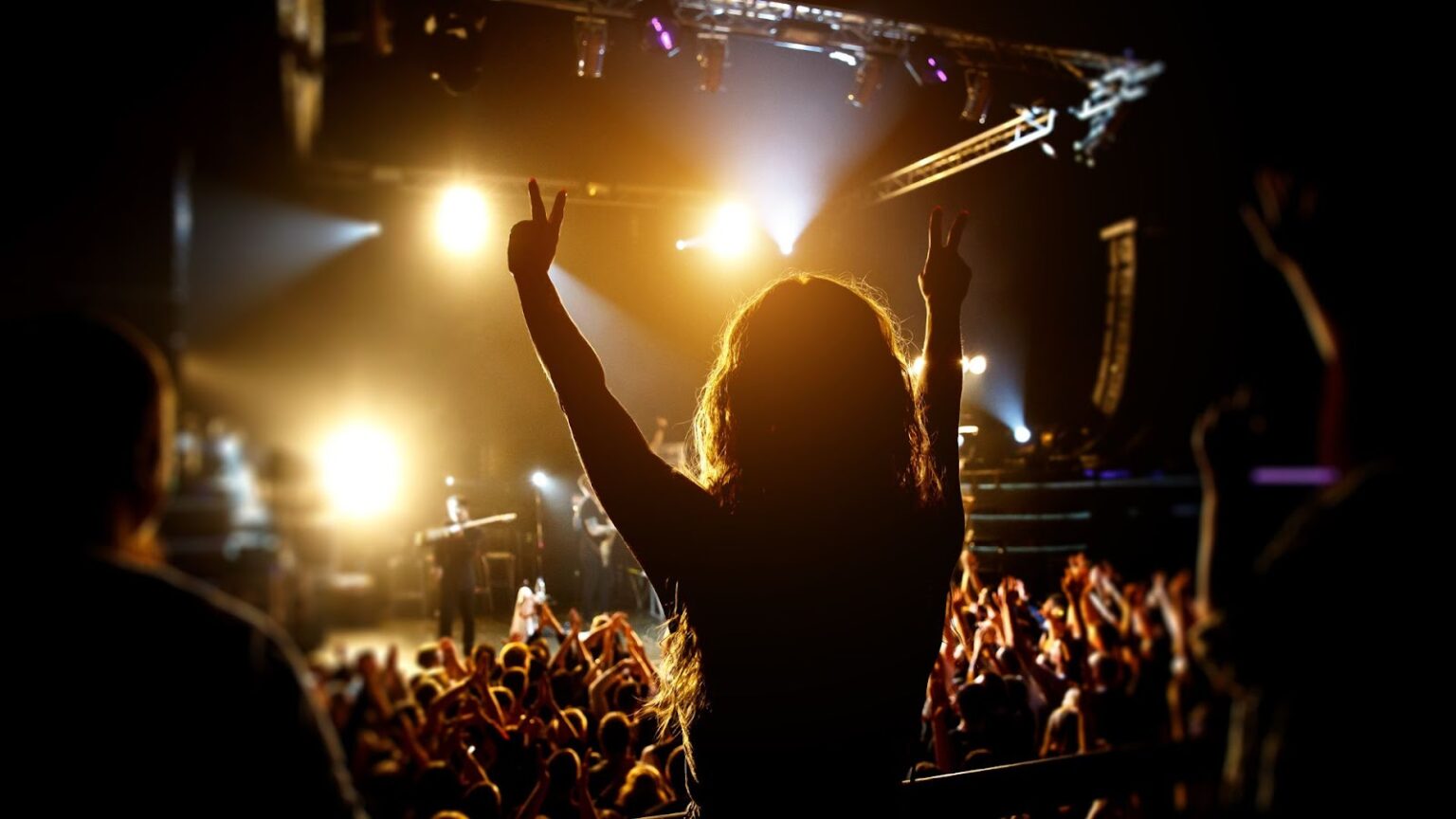 Pessoa com cabelos compridos e ondulados levanta os braços, formando um sinal de paz com cada mão, em meio a uma multidão num show, iluminada pelas luzes do palco. O público ao redor tem os braços levantados e é iluminado por holofotes laranjas.