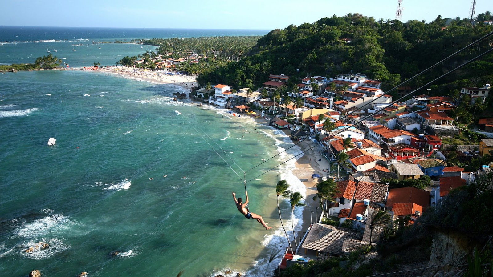 Uma fotografia panorâmica capturada de cima, mostrando uma pessoa praticando tirolesa sobre uma praia tropical. A pessoa está pendurada por um cinto e cabo, deslizando em direção ao mar. Abaixo, há uma pitoresca vila costeira com casas coloridas e telhados de barro, e uma faixa de areia lotada de pessoas. O mar tem tons de azul e verde-água, com ondas suaves.