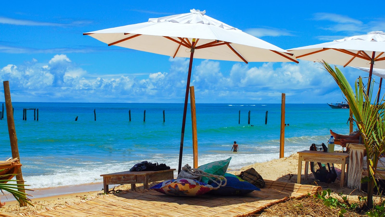 Uma cena de praia tropical em um dia ensolarado. A imagem mostra um espaço de descanso com esteiras de palha no chão e almofadas coloridas, à sombra de grandes guarda-sóis brancos. Há uma espreguiçadeira de madeira e uma mesa na areia. O mar azul-turquesa, com pequenas ondas, se estende até o horizonte, onde barcos flutuam. No mar, há estacas de madeira e uma pessoa tomando banho. O céu é azul e com algumas nuvens brancas.