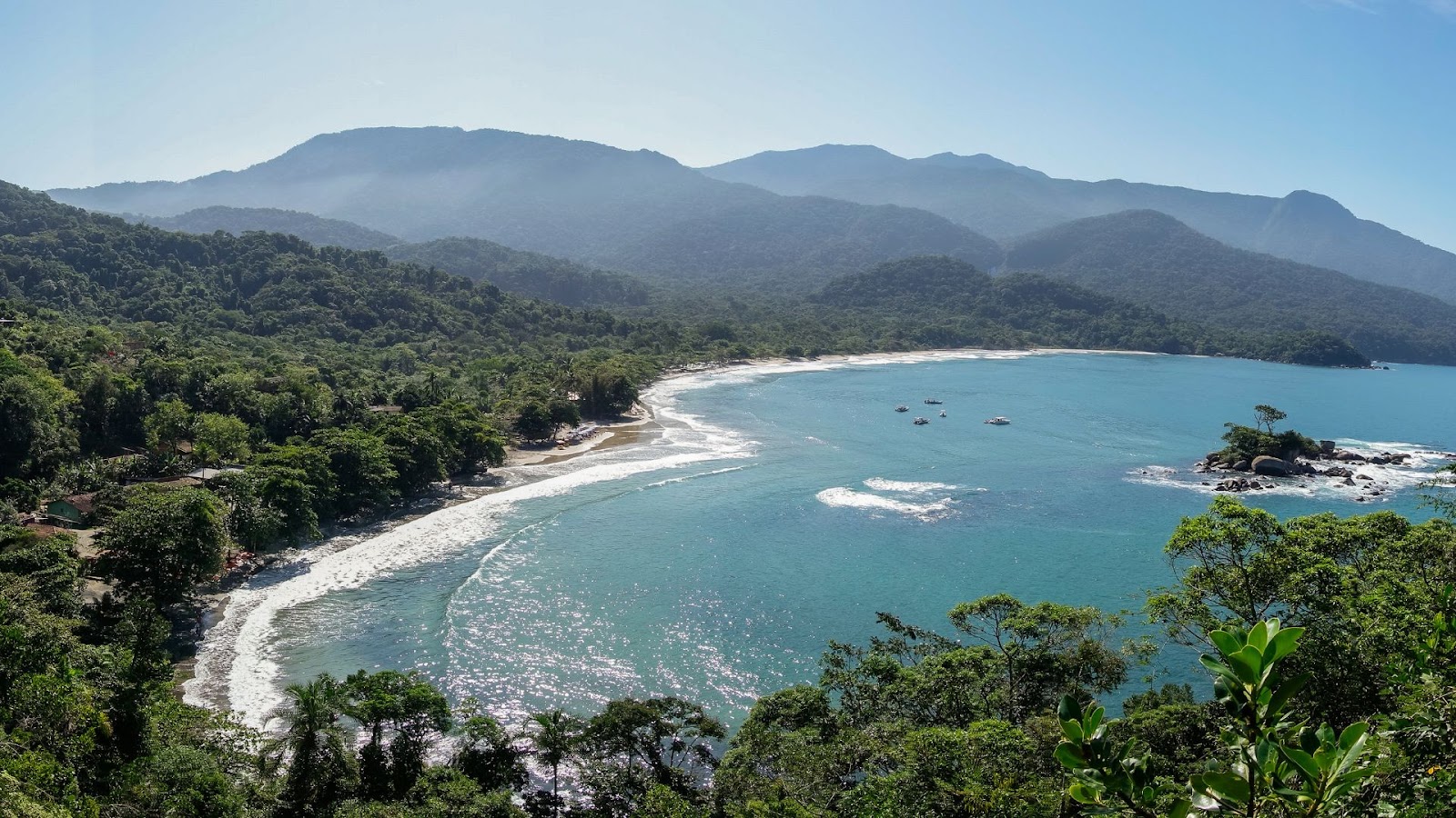 Uma vista aérea de uma baía em forma de meia-lua, com uma praia de areia branca ladeada por floresta tropical densa e montanhas. A água da baía é de um tom de azul-turquesa claro e calmo, com algumas ondas menores quebrando na orla. Há pequenas ilhas rochosas com vegetação espalhadas pela baía e alguns barcos flutuando na água. O céu está limpo, e as montanhas estão envoltas por uma névoa leve.
