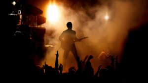 Uma silhueta de um guitarrista no palco, com luzes brilhantes e fumaça ao fundo, enquanto as mãos da multidão se erguem em adoração. A iluminação laranja e amarela cria uma atmosfera energética.