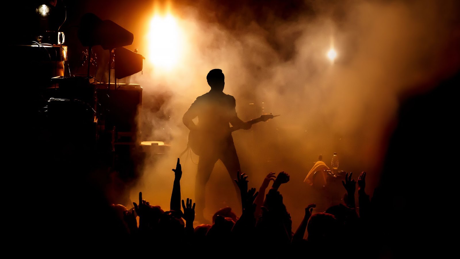 Uma silhueta de um guitarrista no palco, com luzes brilhantes e fumaça ao fundo, enquanto as mãos da multidão se erguem em adoração. A iluminação laranja e amarela cria uma atmosfera energética.
