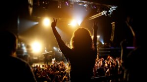 Pessoa com cabelos compridos e ondulados levanta os braços, formando um sinal de paz com cada mão, em meio a uma multidão num show, iluminada pelas luzes do palco. O público ao redor tem os braços levantados e é iluminado por holofotes laranjas.