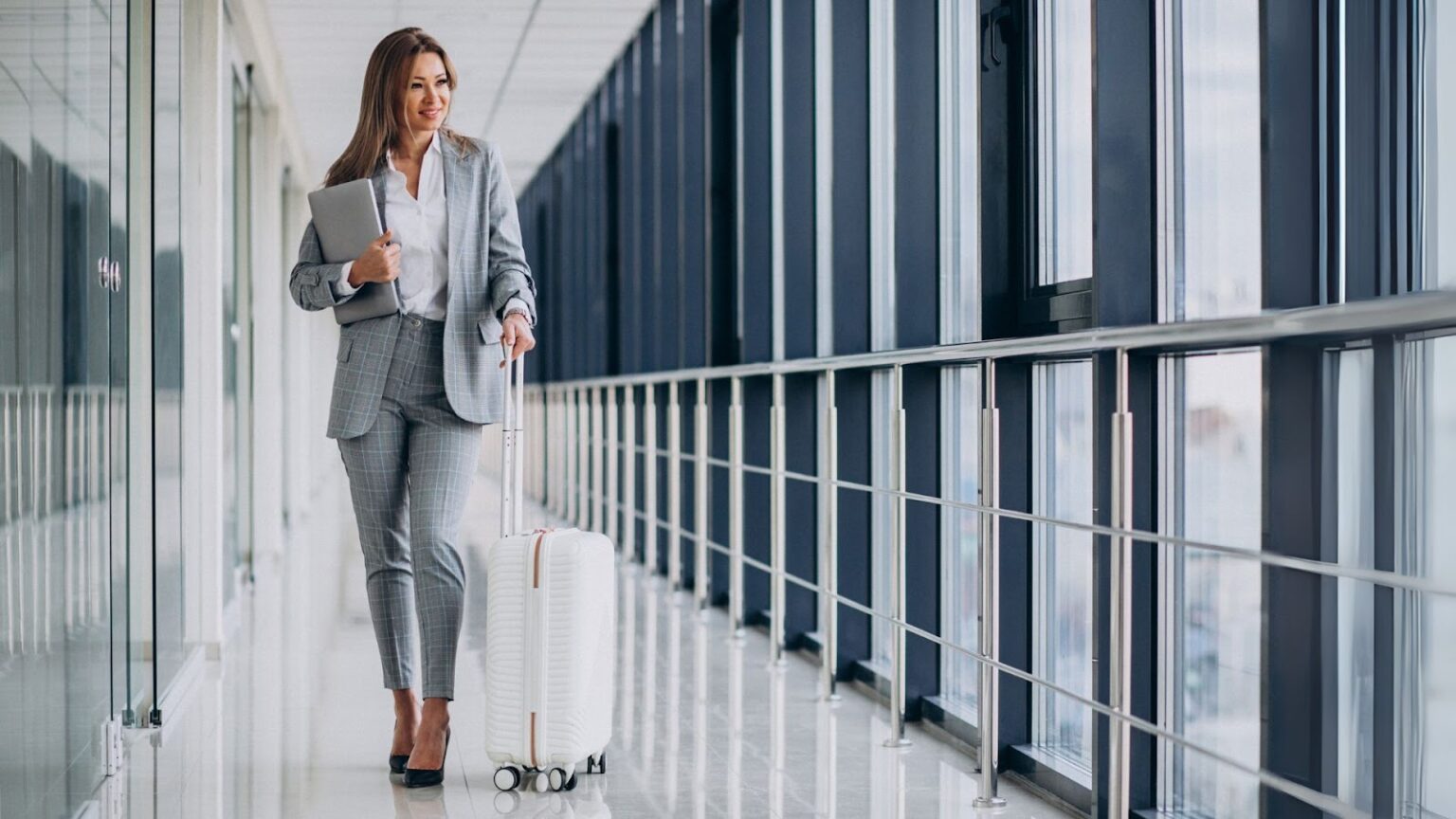 Mulher sorridente em traje de negócios cinza, com um laptop na mão, puxa uma mala de rodinhas branca por um corredor iluminado de aeroporto.