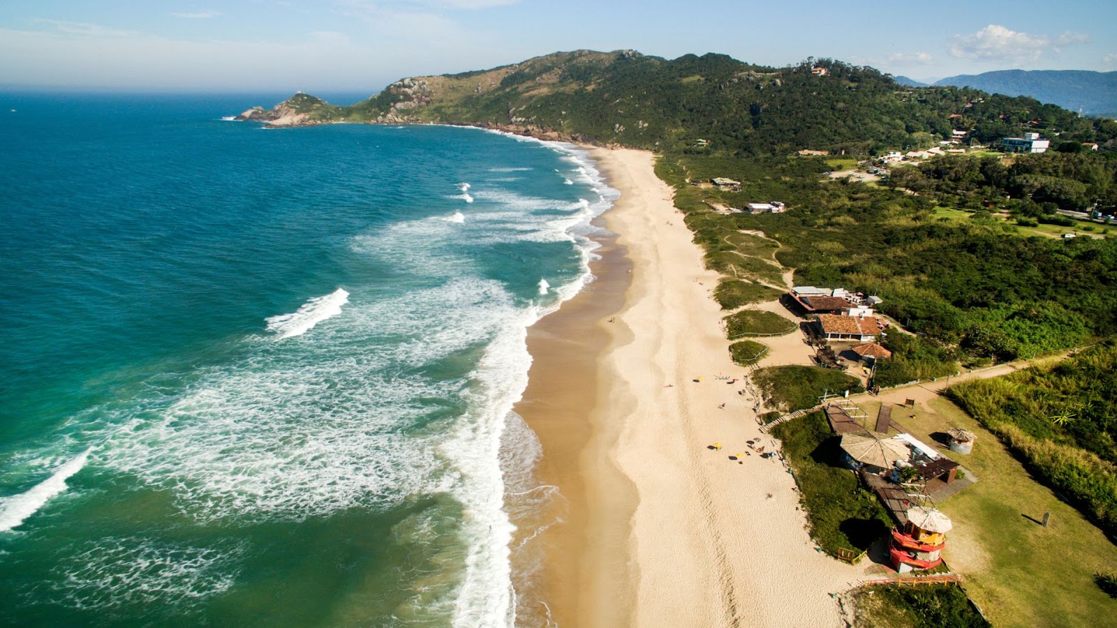 Vista aérea da praia da Lagoinha do Leste, em Florianópolis. A imagem mostra a longa faixa de areia da praia, o mar azul com ondas brancas, uma área verde com vegetação densa e morros ao fundo. Há algumas casas e construções na areia.