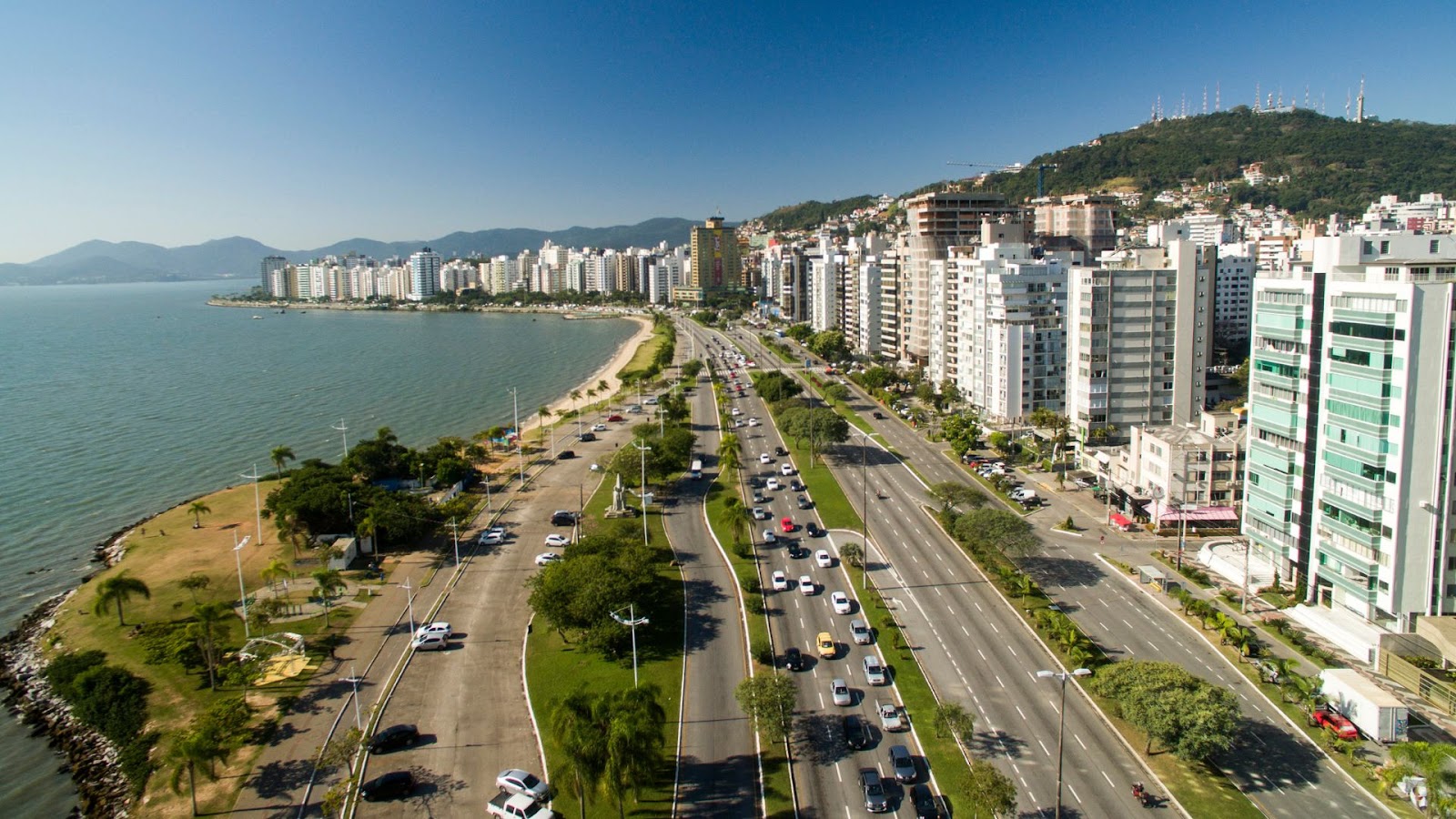 Vista aérea da Beira-Mar Norte, em Florianópolis. A imagem mostra a movimentada avenida com carros, o mar calmo do lado esquerdo e uma orla cheia de árvores e palmeiras. Ao fundo, uma cidade com vários prédios altos e morros. O céu é azul e sem nuvens.