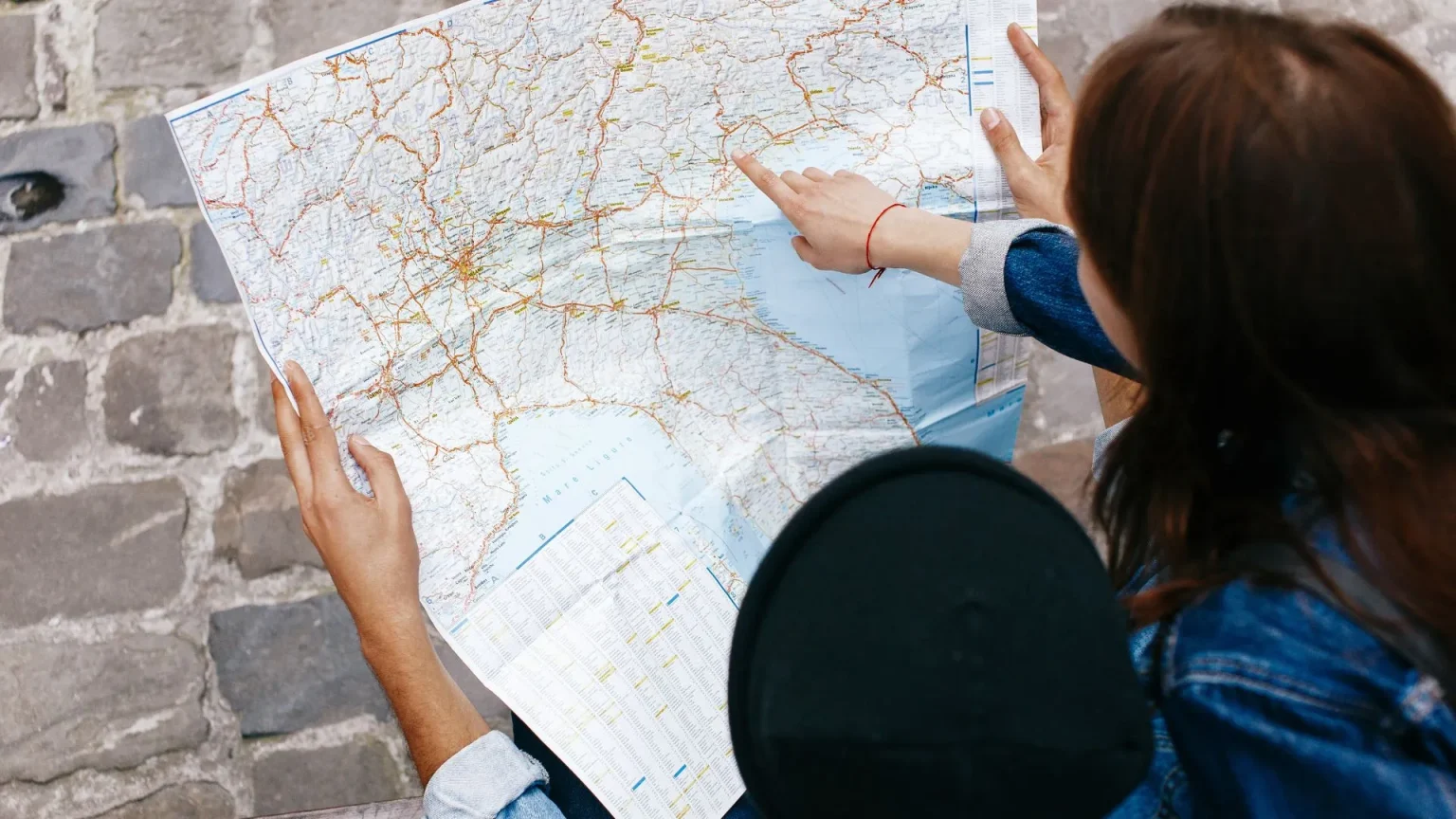 Duas pessoas estão examinando um mapa de papel detalhado. Uma delas, vestindo uma jaqueta jeans, está apontando para uma área específica no mapa, enquanto a outra pessoa segura o mapa. A cena é vista de cima, com o mapa ocupando a maior parte do quadro. As mãos e os braços das pessoas são visíveis, assim como o chão de paralelepípedos. A imagem sugere um momento de planejamento de viagem ou orientação.