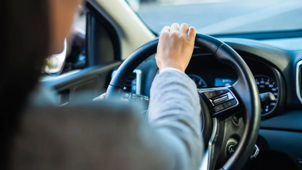 Uma foto em plano médio mostra a parte de cima das costas e o ombro de uma pessoa, vestindo um casaco cinza claro de tricô, sentada ao volante de um carro. A mão direita da pessoa está segurando o volante na parte superior. O volante é preto e possui botões de controle no raio direito. Ao fundo, parcialmente visível, está o painel do carro com o velocímetro. O foco está na mão e no volante.