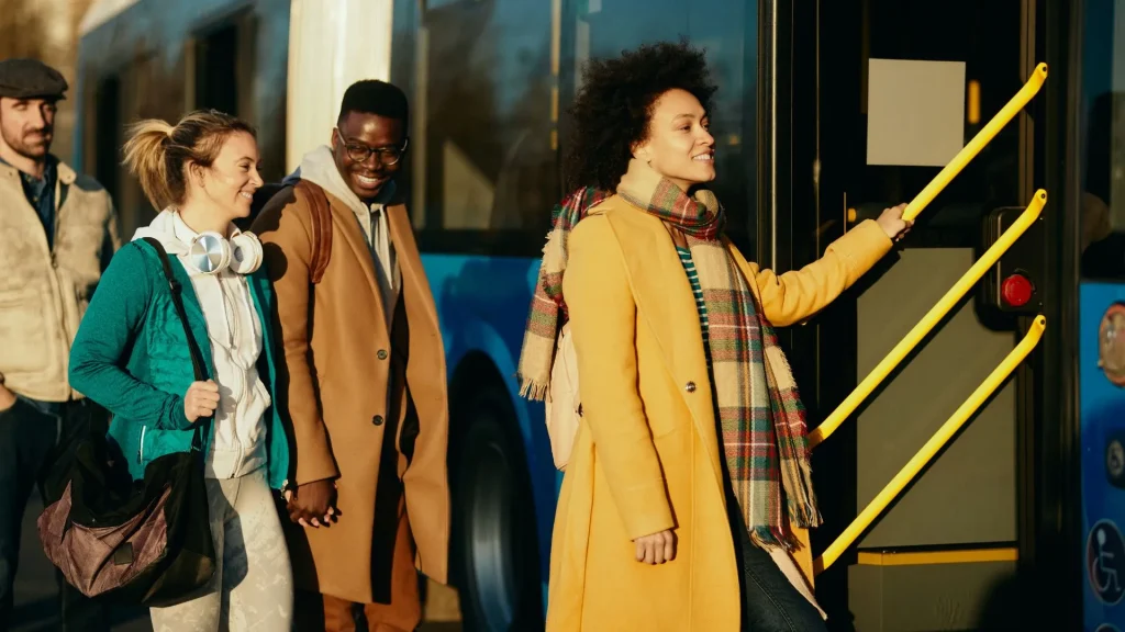 Uma foto externa mostra um grupo de quatro pessoas entrando ou esperando para entrar em um ônibus azul. Uma mulher negra com cabelos crespos, sorrindo e vestindo um casaco amarelo mostarda e um cachecol xadrez, está à direita e prestes a subir os degraus, segurando um corrimão amarelo. À sua esquerda, um homem negro de óculos e um casaco marrom claro está sorrindo. Ao lado dele, uma mulher branca loira, de fone de ouvido no pescoço e casaco verde, sorri para ele. Um quarto homem, de boné, está parcialmente visível mais à esquerda. A luz do sol atinge o grupo.