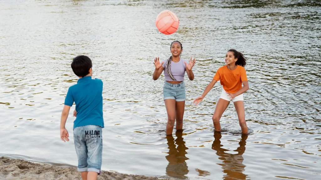 Três crianças brincam em um rio. Duas meninas, de pé na água até a altura das canelas, estão no centro e na direita da imagem, e um menino, de pé na areia, está de costas para o observador na parte esquerda. A menina no centro, que veste uma blusa roxa, joga uma bola rosa para o alto, em direção ao menino. A segunda menina, de blusa laranja, está à direita e parece pronta para a jogada. O rio tem uma superfície com pequenas ondulações. O fundo da imagem está desfocado, mostrando a água em tom acinzentado.