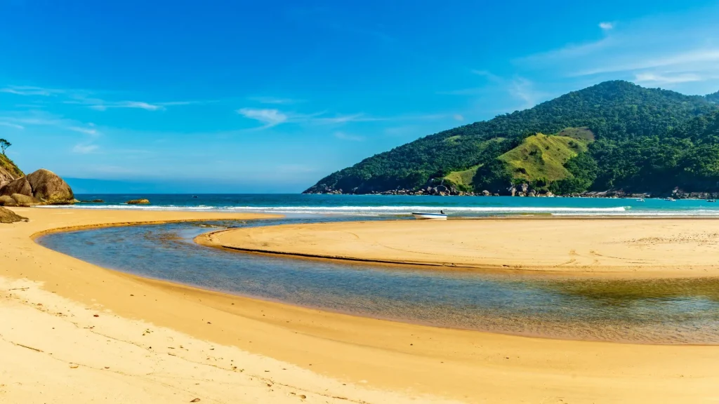 A imagem mostra uma paisagem de praia deslumbrante, com um rio de águas calmas que desagua no mar. A areia dourada e a água azul-clara dominam a cena. Ao fundo, uma imponente montanha coberta por mata verde exuberante se destaca contra o céu azul sem nuvens. Uma pequena embarcação branca está ancorada perto da foz do rio.