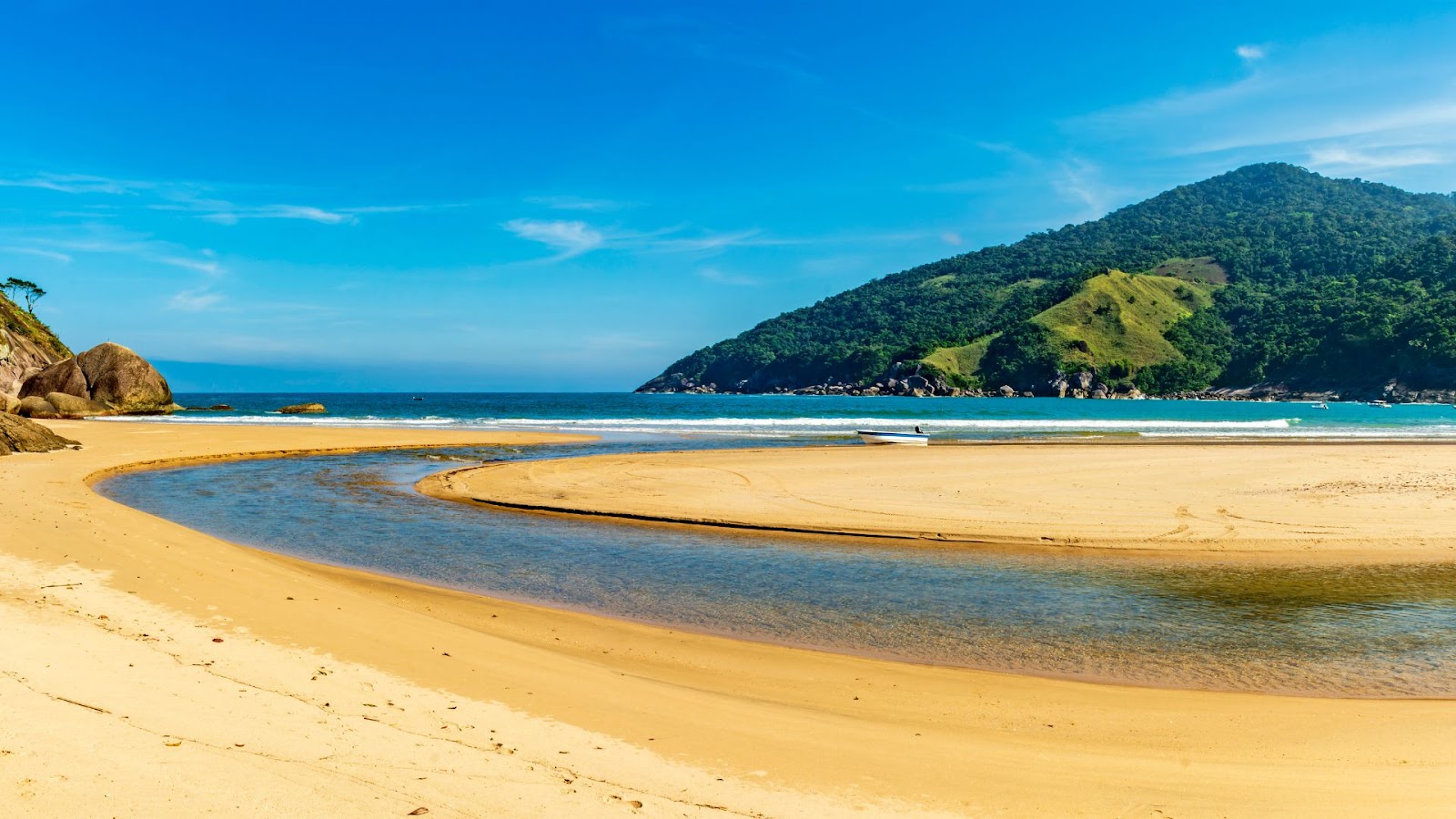 A imagem mostra uma paisagem de praia deslumbrante, com um rio de águas calmas que desagua no mar. A areia dourada e a água azul-clara dominam a cena. Ao fundo, uma imponente montanha coberta por mata verde exuberante se destaca contra o céu azul sem nuvens. Uma pequena embarcação branca está ancorada perto da foz do rio.