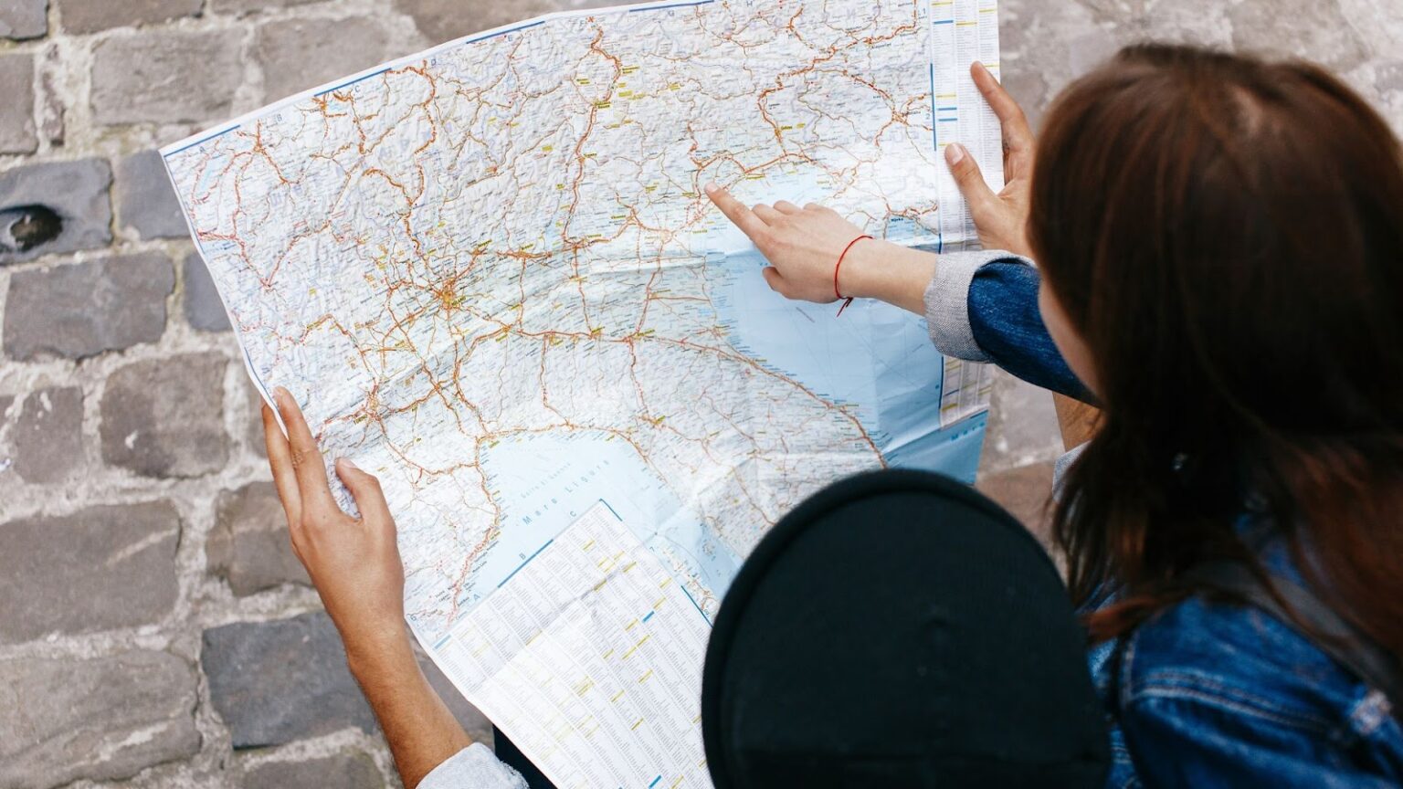 Duas pessoas estão examinando um mapa de papel detalhado. Uma delas, vestindo uma jaqueta jeans, está apontando para uma área específica no mapa, enquanto a outra pessoa segura o mapa. A cena é vista de cima, com o mapa ocupando a maior parte do quadro. As mãos e os braços das pessoas são visíveis, assim como o chão de paralelepípedos. A imagem sugere um momento de planejamento de viagem ou orientação.