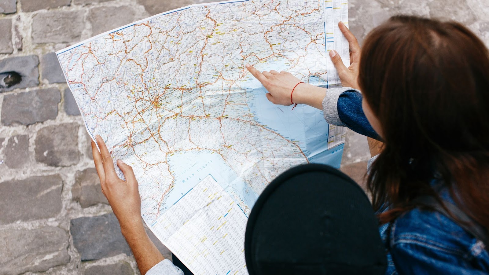 Duas pessoas estão examinando um mapa de papel detalhado. Uma delas, vestindo uma jaqueta jeans, está apontando para uma área específica no mapa, enquanto a outra pessoa segura o mapa. A cena é vista de cima, com o mapa ocupando a maior parte do quadro. As mãos e os braços das pessoas são visíveis, assim como o chão de paralelepípedos. A imagem sugere um momento de planejamento de viagem ou orientação.