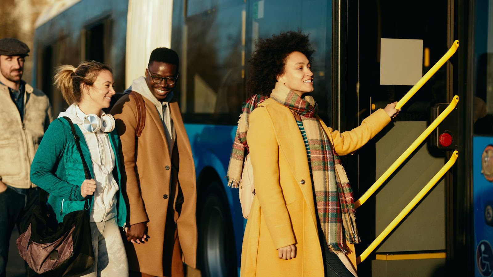 Uma foto externa mostra um grupo de quatro pessoas entrando ou esperando para entrar em um ônibus azul. Uma mulher negra com cabelos crespos, sorrindo e vestindo um casaco amarelo mostarda e um cachecol xadrez, está à direita e prestes a subir os degraus, segurando um corrimão amarelo. À sua esquerda, um homem negro de óculos e um casaco marrom claro está sorrindo. Ao lado dele, uma mulher branca loira, de fone de ouvido no pescoço e casaco verde, sorri para ele. Um quarto homem, de boné, está parcialmente visível mais à esquerda. A luz do sol atinge o grupo.