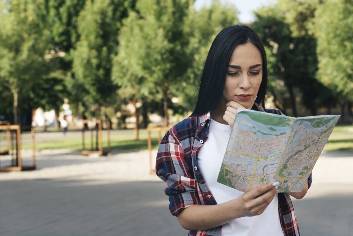 Uma foto externa mostra uma jovem mulher de cabelos escuros longos no centro, usando uma camiseta branca e uma camisa xadrez por cima, segurando e examinando um mapa dobrado. Ela tem uma expressão concentrada, com a mão direita no queixo. O fundo é um caminho pavimentado em um parque ou área verde, com árvores frondosas. A luz do sol está filtrada pelas árvores.