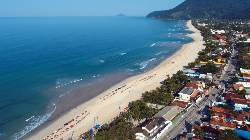 Vista aérea de uma praia curva com mar verde-água e ondas suaves. Em primeiro plano, uma encosta com vegetação densa. Ao fundo, prédios baixos, areia clara e morros cobertos por mata atlântica sob um céu azul claro.