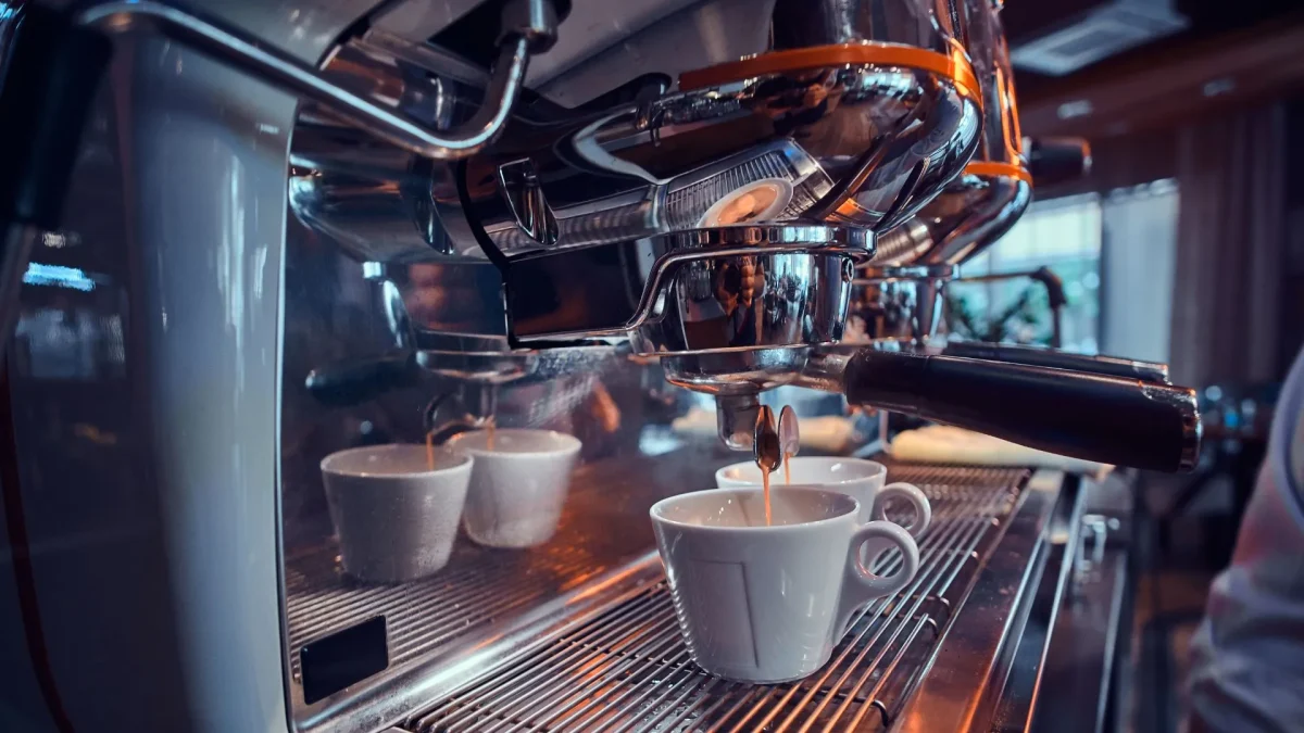 Um close-up em uma máquina de café expresso brilhante de cafeteria, mostrando a bandeja de metal e o cabeçote (grupo). Uma xícara de cerâmica branca está posicionada sob o bico de extração, com outras duas xícaras ao fundo, prontas para o preparo do café.