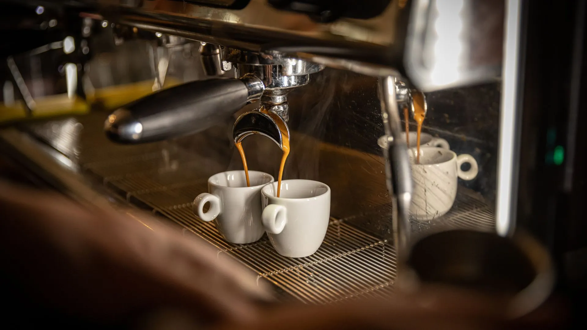 Uma foto em close-up, com iluminação quente e baixa, mostra uma máquina de café espresso profissional em ação. O café recém-tirado escorre em jatos duplos de um porta-filtro (alça preta à esquerda) diretamente para duas pequenas xícaras de cerâmica branca. O vapor sobe levemente da máquina e da bebida. Um detalhe de uma mão aparece no canto inferior esquerdo, manuseando a máquina.
