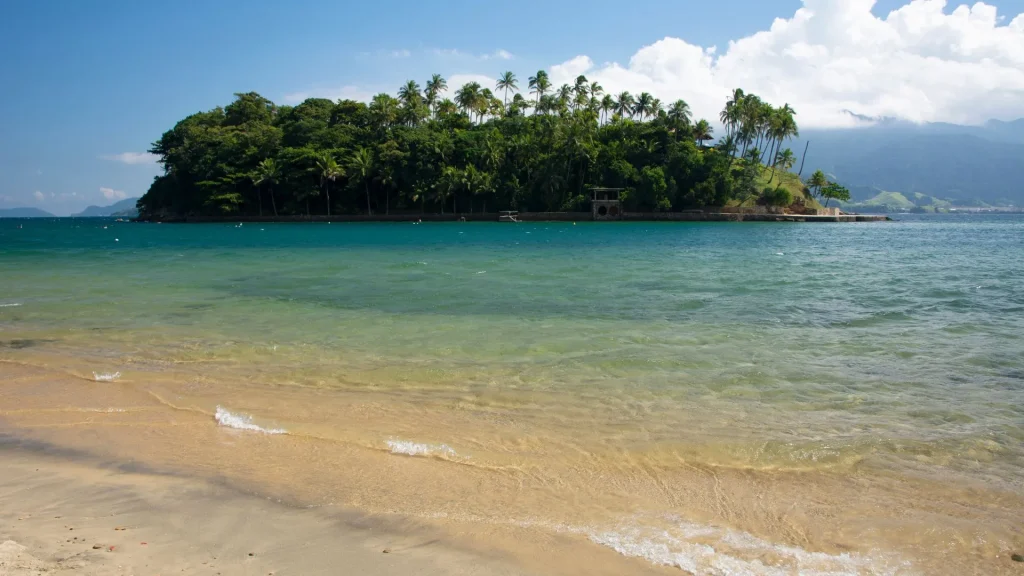 Vista de uma praia tropical com areia clara, água do mar transparente em tons de verde e azul, e uma pequena ilha coberta por densa vegetação e coqueiros (palmeiras) em segundo plano. O céu está parcialmente nublado, e montanhas são visíveis ao fundo, à direita.