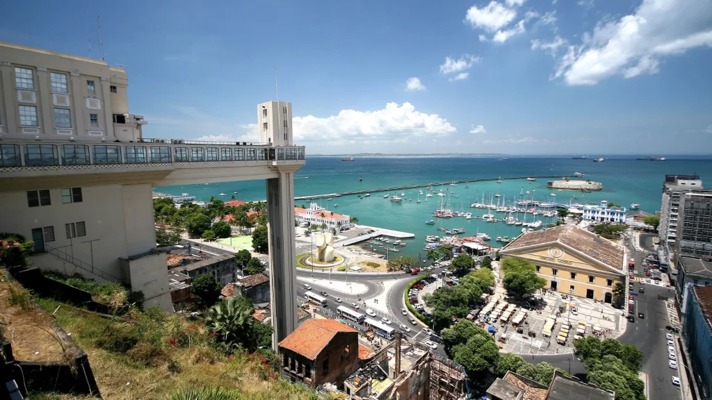 Vista panorâmica de Salvador, Bahia. Em primeiro plano, o Elevador Lacerda, uma torre de concreto branca que conecta os bairros inferior e superior. Abaixo, vê-se um movimentado cruzamento e edifícios históricos. Ao fundo, a Baía de Todos os Santos está repleta de barcos e navios, estendendo-se até o horizonte sob um céu azul claro com nuvens esparsas.