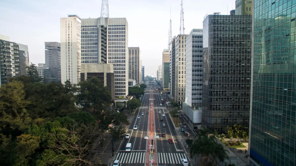 Vista em plano aberto da Avenida Paulista, em São Paulo, com um canteiro central. A avenida está cercada por arranha-céus altos e modernos de ambos os lados. Árvores e vegetação preenchem a parte inferior e as margens da rua, com tráfego de carros em ambas as direções. O céu está claro e um pouco encoberto.