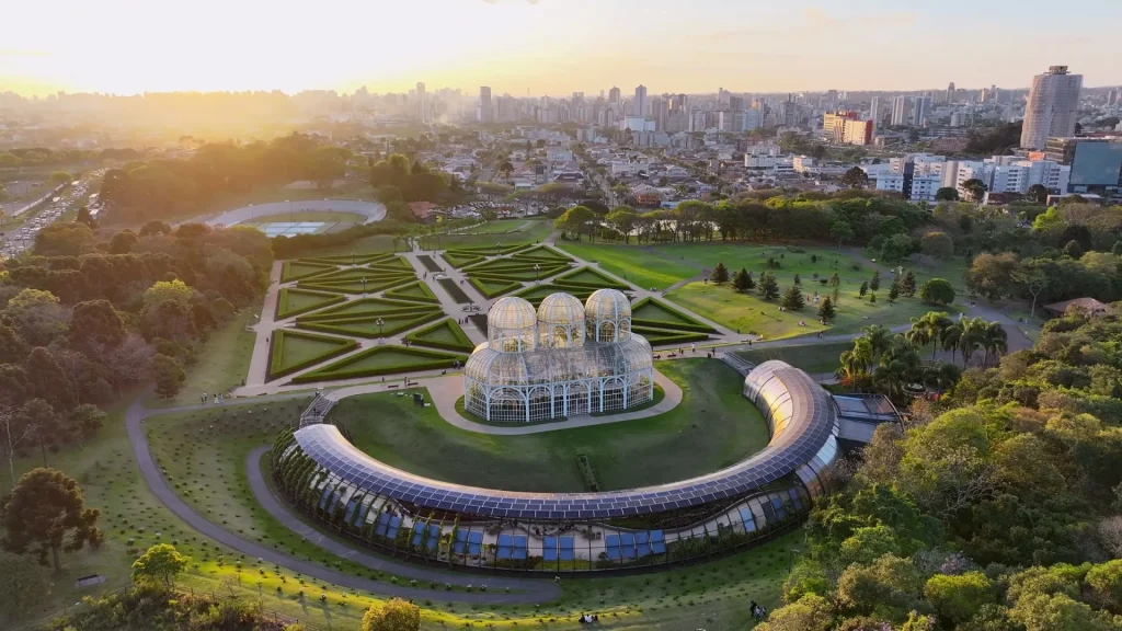 Vista aérea do Jardim Botânico de Curitiba, Paraná. Em destaque, a icônica estufa de vidro Art Nouveau, cercada por jardins simétricos formais no estilo francês. Um edifício semicircular coberto por painéis solares se curva em torno da estufa. A área verde se estende até o fundo, onde o horizonte da cidade, cheio de arranha-céus, é visível sob o brilho quente do pôr do sol ou nascer do sol.