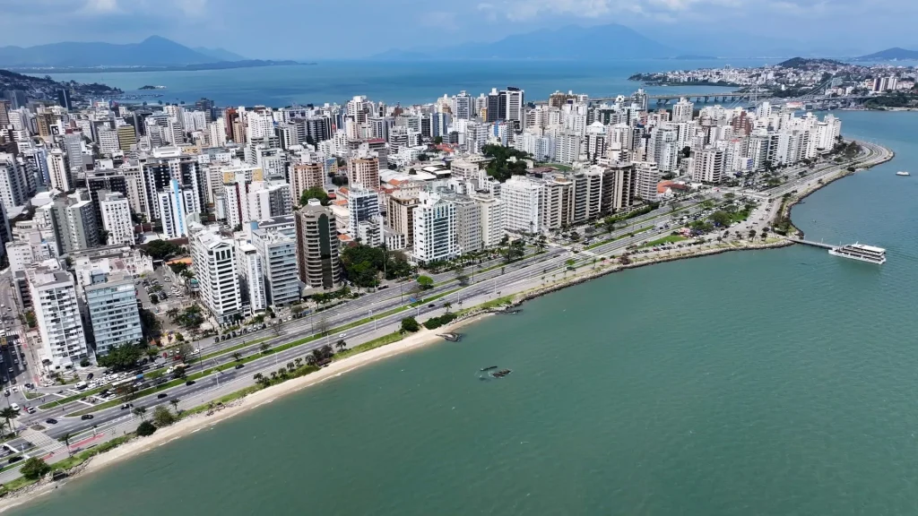 Vista aérea do horizonte de Florianópolis, SC, mostrando a orla da Avenida Beira-Mar Norte e uma densa área de edifícios residenciais e comerciais junto à praia. O mar verde-azulado está em primeiro plano, com montanhas e o continente visíveis ao fundo, e a Ponte Hercílio Luz parcialmente visível no canto superior direito.