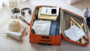 Mala de viagem de cor laranja aberta, recheada com roupas dobradas (shorts brancos, jeans) e itens pessoais, como um caderno ou bloco de anotações, uma caneta e um celular. Ao redor da mala, sobre um piso claro, estão dispostos outros acessórios essenciais para viagem: um chapéu de palha de aba larga, um secador de cabelo, uma escova de cabelo, fones de ouvido, uma câmera fotográfica e um pequeno recipiente de metal. No lado direito da mala, toalhas dobradas, cabides de madeira e frascos de amenities (sabonete líquido, shampoo, etc.) completam a cena.