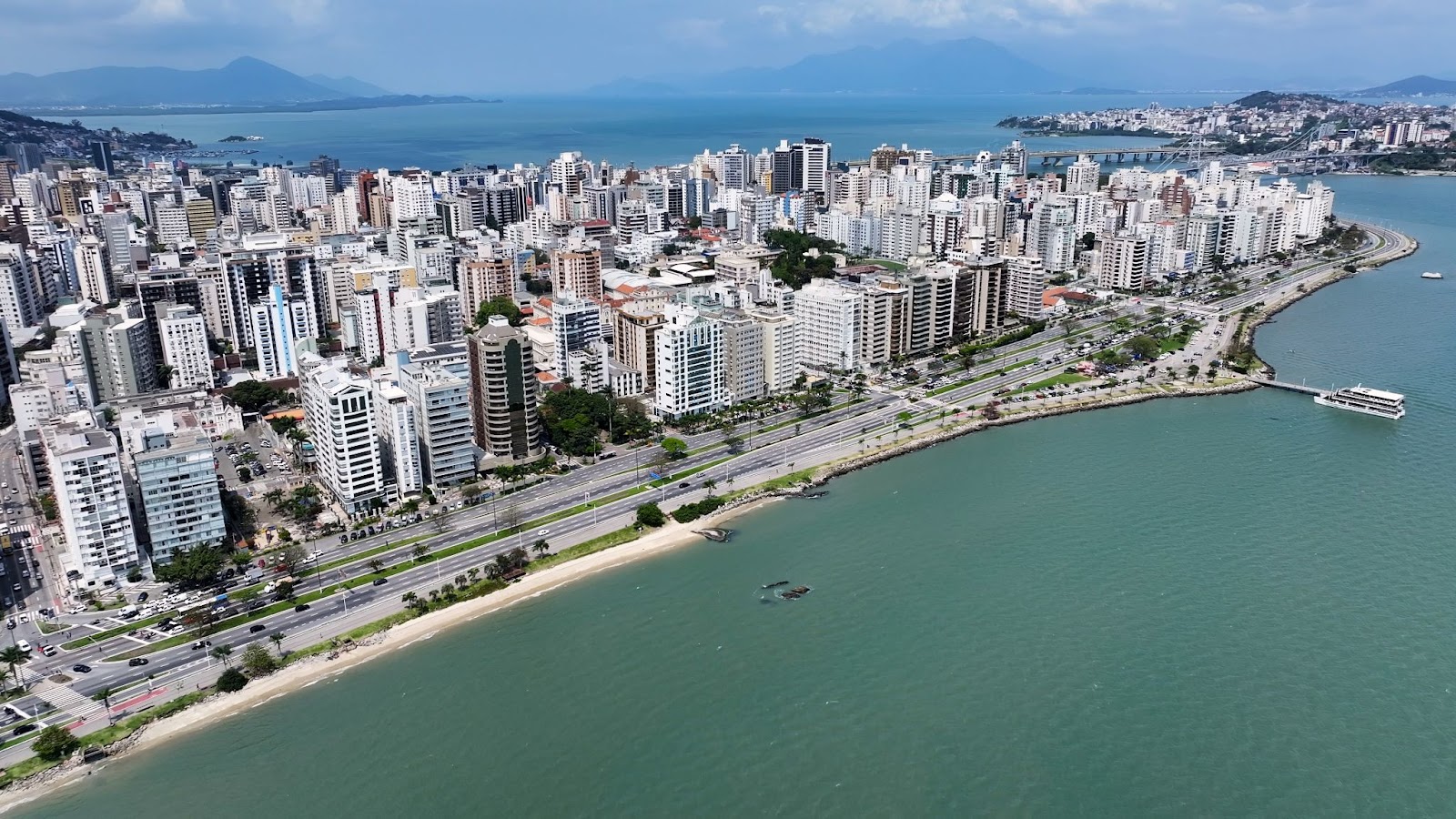 Vista aérea do horizonte de Florianópolis, SC, mostrando a orla da Avenida Beira-Mar Norte e uma densa área de edifícios residenciais e comerciais junto à praia. O mar verde-azulado está em primeiro plano, com montanhas e o continente visíveis ao fundo, e a Ponte Hercílio Luz parcialmente visível no canto superior direito.