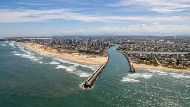 Vista aérea panorâmica e em um dia ensolarado da orla de uma cidade litorânea (identificada como Torres, Rio Grande do Sul), com destaque para o local onde um rio encontra o mar.