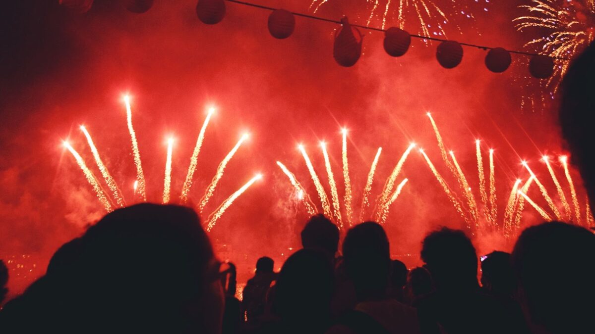 Exibição noturna intensa de fogos de artifício em tons de vermelho e dourado, com algumas explosões em formato de palmeira. A cena é vista por trás de uma multidão silhuetada, com balões de papel pendurados em um cordão sobre as cabeças das pessoas, criando um ambiente de celebração.