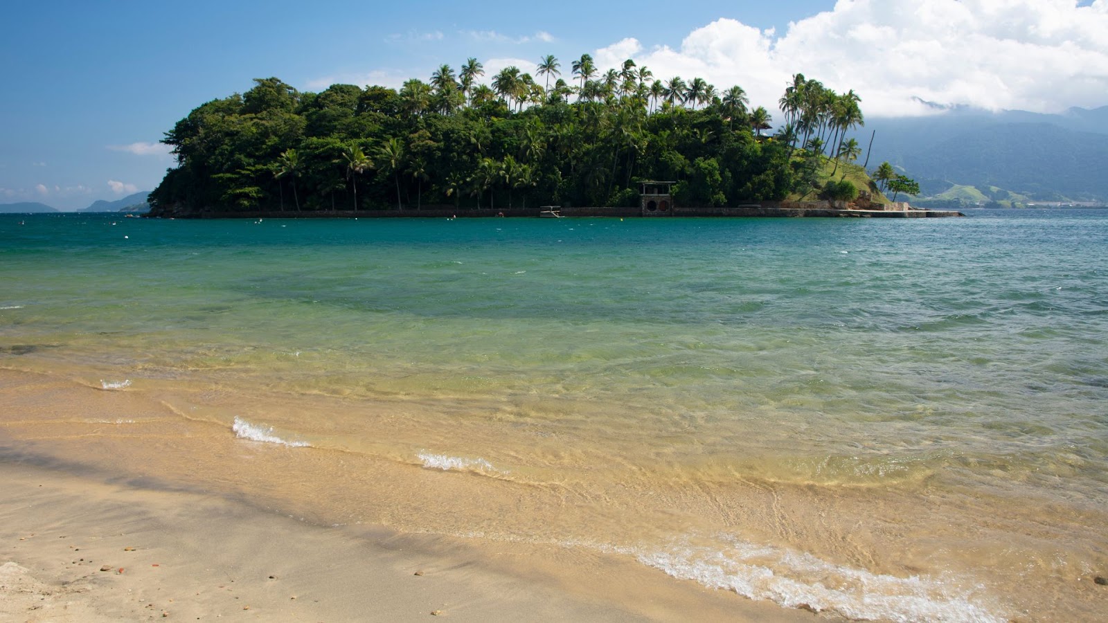 Ilhabela Vista de uma praia tropical com areia clara, água do mar transparente em tons de verde e azul, e uma pequena ilha coberta por densa vegetação e coqueiros (palmeiras) em segundo plano. O céu está parcialmente nublado, e montanhas são visíveis ao fundo, à direita.