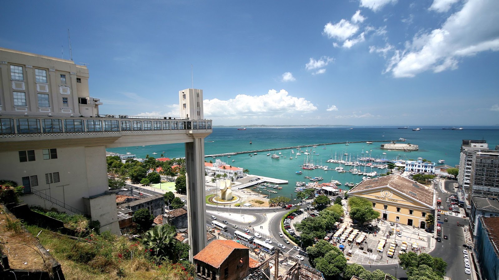 Salvador Vista panorâmica de Salvador, Bahia. Em primeiro plano, o Elevador Lacerda, uma torre de concreto branca que conecta os bairros inferior e superior. Abaixo, vê-se um movimentado cruzamento e edifícios históricos. Ao fundo, a Baía de Todos os Santos está repleta de barcos e navios, estendendo-se até o horizonte sob um céu azul claro com nuvens esparsas.