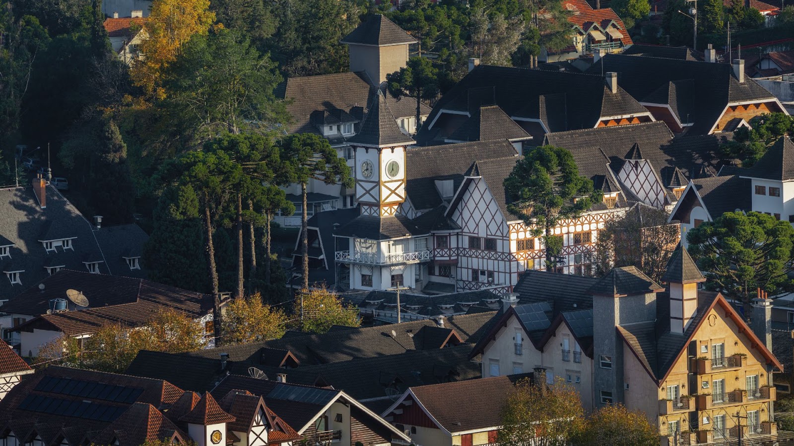 Campos do Jordão Vista aérea de uma vila ou bairro com forte influência de arquitetura alpina, no estilo enxaimel. O edifício central é branco e marrom-escuro, com uma torre de relógio proeminente. O telhado escuro dos edifícios se estende em cascata pela colina, cercado por densas árvores de pinheiros (araucárias) e alguma folhagem de outono, sugerindo um clima frio.