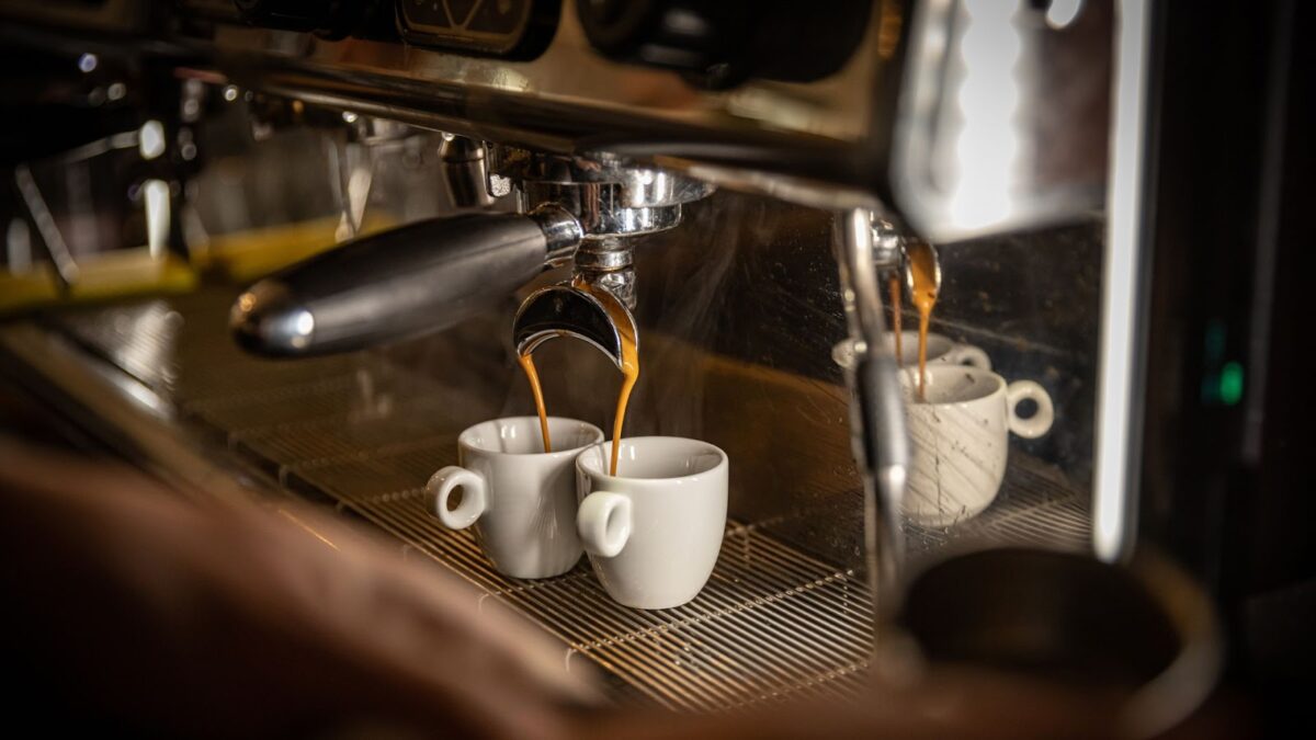 Uma foto em close-up, com iluminação quente e baixa, mostra uma máquina de café espresso profissional em ação. O café recém-tirado escorre em jatos duplos de um porta-filtro (alça preta à esquerda) diretamente para duas pequenas xícaras de cerâmica branca. O vapor sobe levemente da máquina e da bebida. Um detalhe de uma mão aparece no canto inferior esquerdo, manuseando a máquina.