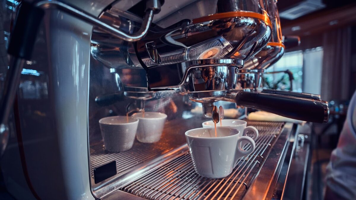 Um close-up em uma máquina de café expresso brilhante de cafeteria, mostrando a bandeja de metal e o cabeçote (grupo). Uma xícara de cerâmica branca está posicionada sob o bico de extração, com outras duas xícaras ao fundo, prontas para o preparo do café.